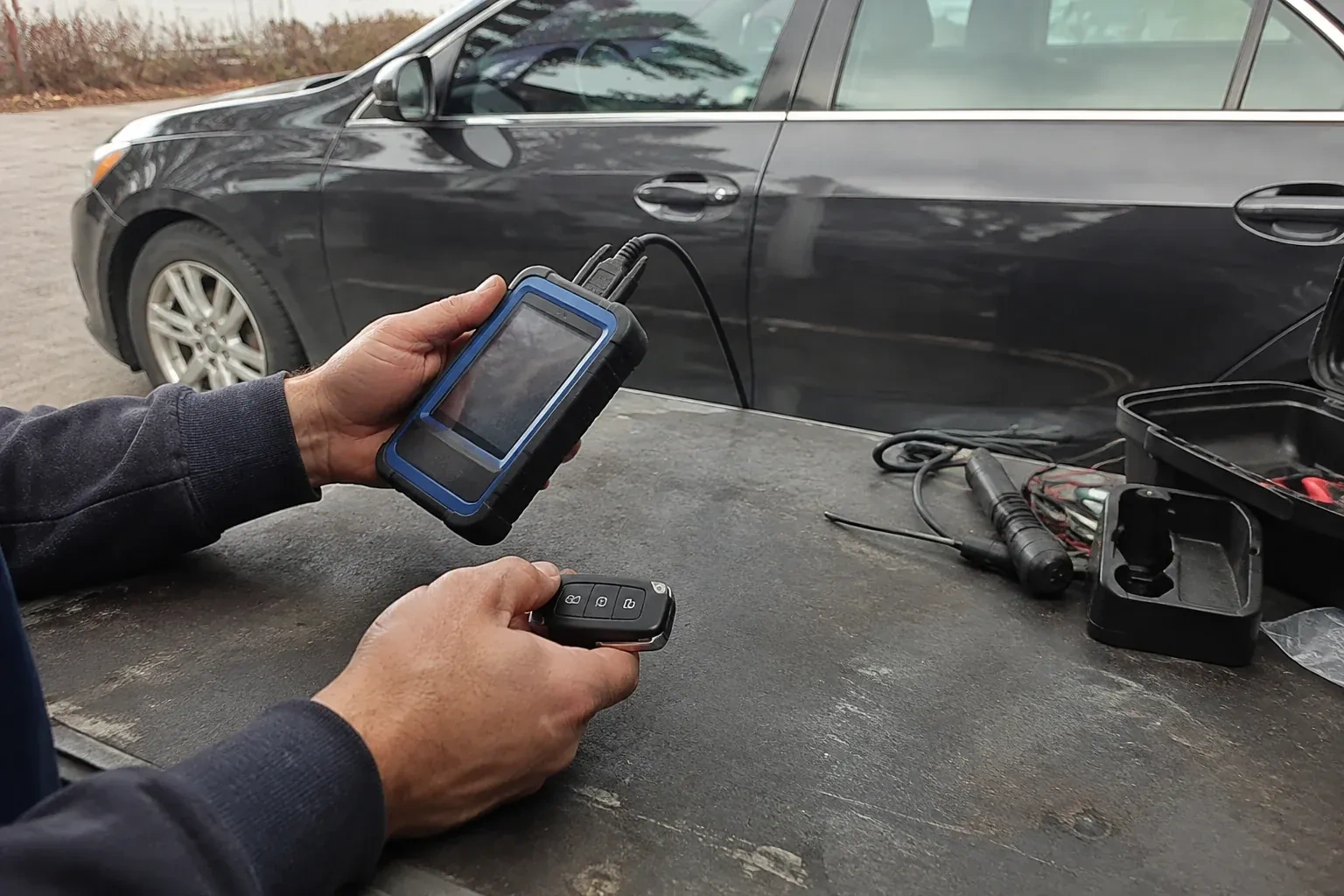 Hands holding a blue-edged electronic device and car-key fob at a worktable beside a parked car. — Transponder Key Programming, Collingwood
