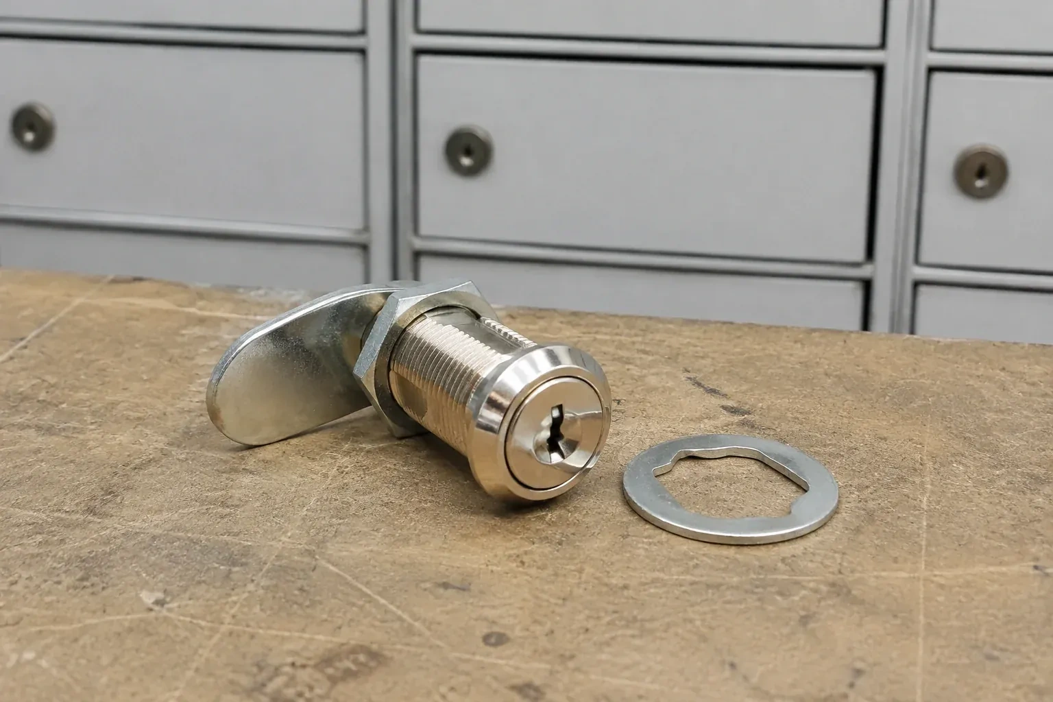 Detached threaded cam lock and mounting washer on a workbench in front of closed mail compartments. — Mailbox Lock Replacement, Collingwood